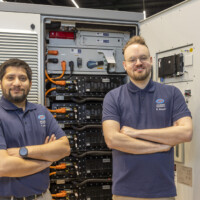 Two JST Power Equipment engineers pose in front of an open battery energy storage cabinet, revealing modular lithium-ion battery packs and internal power electronics. Two JST Power Equipment engineers standing confidently in front of an open battery energy storage cabinet at Ennovation 2025, Messezentrum Salzburg.