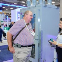 Two attendees standing beside an Outdoor substation breaker at JST Power Equipment’s DISTRIBUTECH 2025 booth