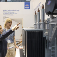 Visitors engage with a detailed transformer model at the JST Power Equipment booth, Smarter E Europe 2025, Messezentrum München. Two women discussing a high-voltage power transformer model at the JST Power Equipment booth during Smarter E Europe 2025 in Munich.