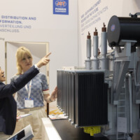 Visitors engage with a detailed small power transformer model at the JST Power Equipment booth at Smarter E Europe 2025, Messezentrum München. Two women discussing a high-voltage power transformer model at the JST Power Equipment booth during Smarter E Europe 2025 in Munich.