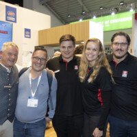JST team and partners enjoying the vibrant atmosphere at Smarter E Europe 2025, celebrating new collaborations and innovations. Group of five smiling people at an exhibition booth during Smarter E Europe 2025, posing together with drinks in hand and wearing event badges.