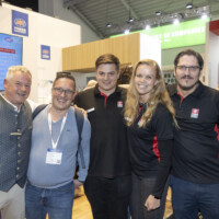 JST team and partners enjoying the vibrant atmosphere at Smarter E Europe 2025, celebrating new collaborations and innovations. Group of five smiling people at an exhibition booth during Smarter E Europe 2025, posing together with drinks in hand and wearing event badges.