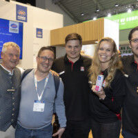 JST team and partners enjoying the vibrant atmosphere at the trade show, celebrating new collaborations and innovations. Group of five smiling people at an exhibition booth, posing together with drinks in hand and wearing event badges.