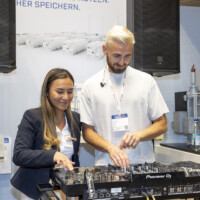 A JST Power member joins the DJ at the JST Power Equipment booth for a fun, interactive moment during Smarter E Europe 2025 in Messezentrum München. Female attendee and DJ interacting at a Pioneer DJ setup at the JST Power Equipment booth during Smarter E Europe 2025 in Munich.