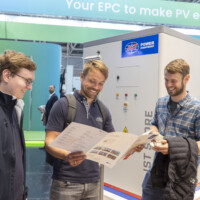 Enthusiastic attendees dive into JST Power’s energy storage offerings at The Smarter E Europe 2025, engaging with informative product materials. Three men smiling and looking at a brochure in front of a JST Power energy storage unit at a trade show booth.