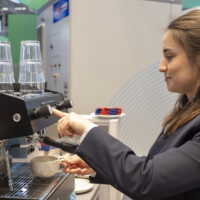 A JST Power team member prepares coffee for booth visitors at The Smarter E Europe 2025, combining hospitality with innovation. A woman in a dark blazer operating a coffee machine, brewing a fresh cup of coffee at an exhibition booth with a man in the background.